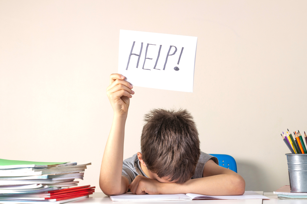 frustrated child with head down on desk holding a help sign struggling with a book that is too hard